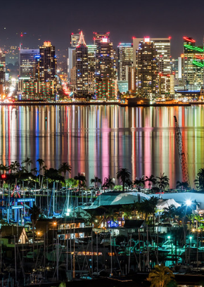 San Diego cityscape waterfront view at night with docked boats in the foreground, streaks of multicolored reflections of the city building lights on the water of the bay, with brightly lit tall city buildings in the distance.